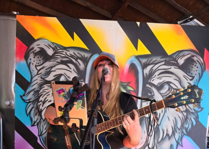 Female musician performing live with an acoustic guitar in front of a colorful bear mural at Contentment Brewing.