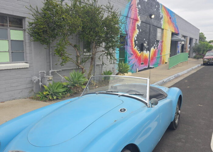 Vintage light blue convertible parked outside a mural-covered brewery wall featuring a tie-dye hop design.